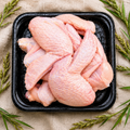 Raw chicken wings in a black plastic tray, arranged on a beige fabric background with green sprigs around the edges.
