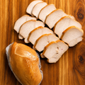 Sliced chicken arranged on a wooden cutting board next to a whole raw chicken breast wrapped in plastic.